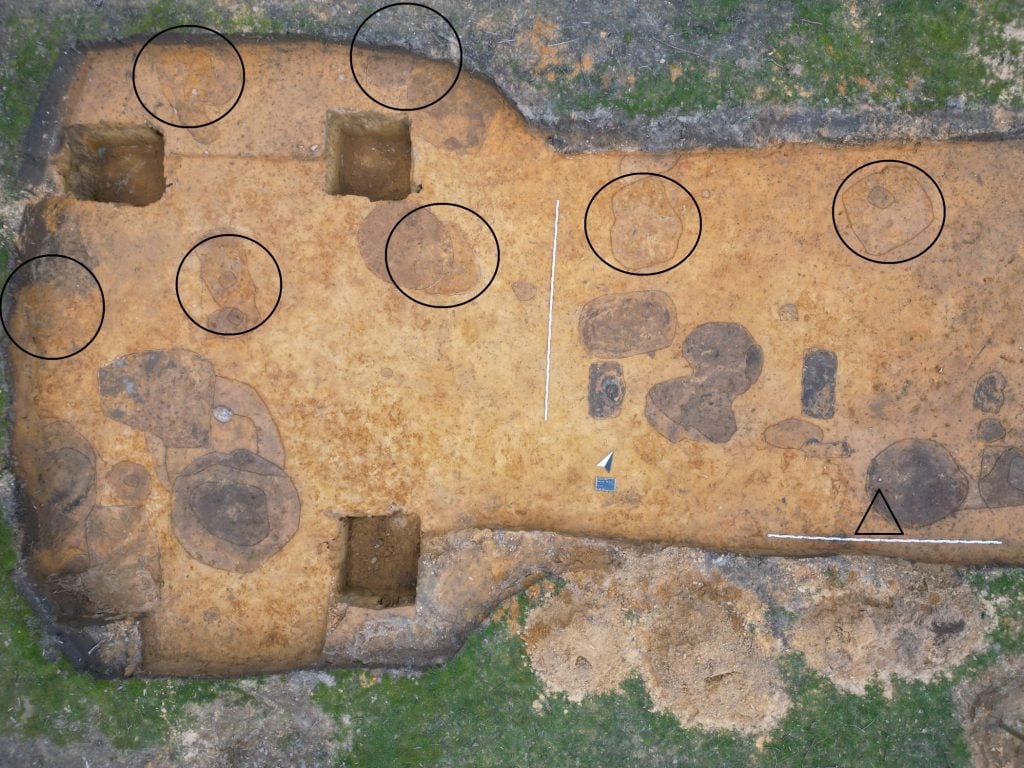 Aa aerial photograph of a large brown excavation site surrounded by green grass and punctuated with circles and arrows marking different points of interest.