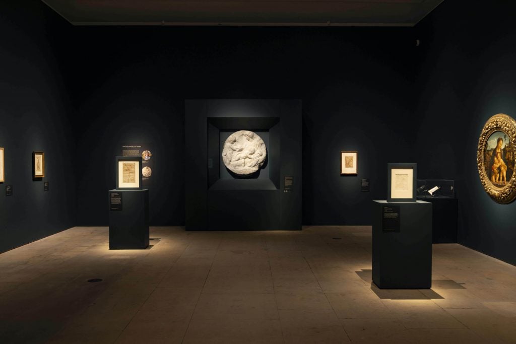 View of a dark gallery with several plinths lit from above showing Renaissance drawings and a large circular sculpture on the back wall.