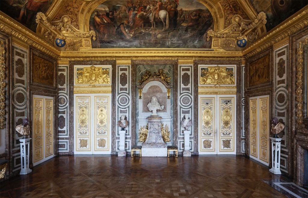 A photograph of Le Salon de Diane at Versailles, featuring a white bust of Louis XIV beneath an ornate celing and between ornate wall decorations.