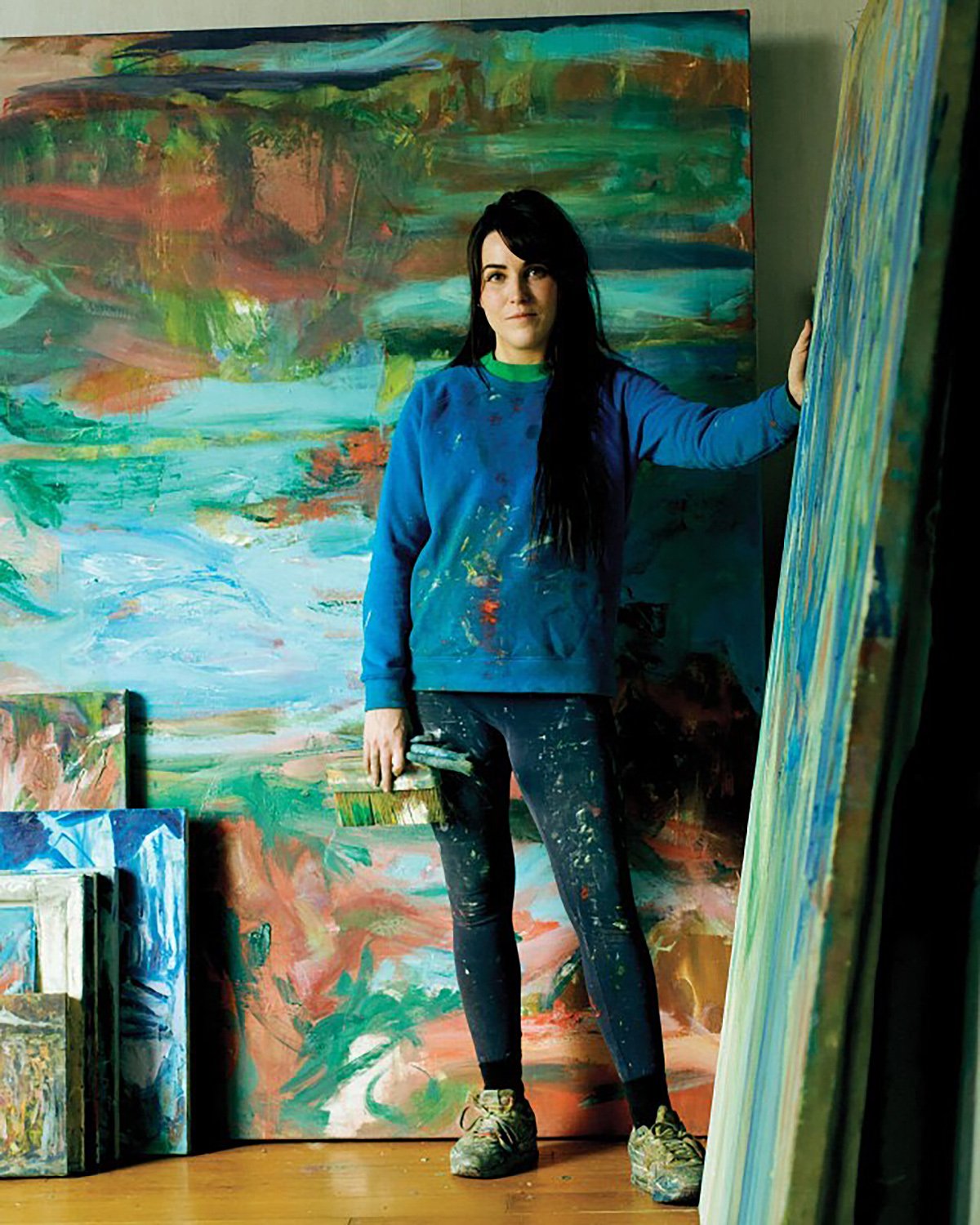 A woman in a paint-splattered sweatshirt standing in a studio surrounded by paintings