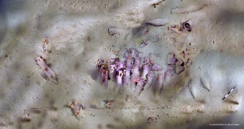 An underwater photograph taken above a rows of purple-tinged ceramic vessels, many of which are slightly covered with sand amongst the ocean floor off the coast of Sicily.