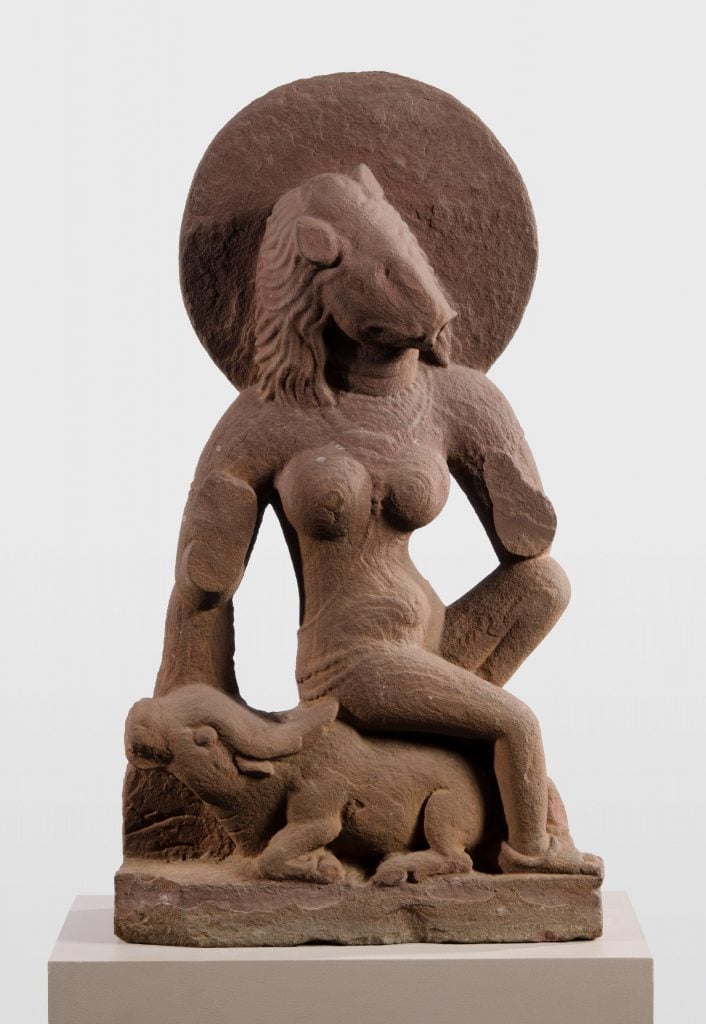 small brown statue on a pedestal of a humanoid creature with a sow for a head with large breasts seated in front of a dog looking creature