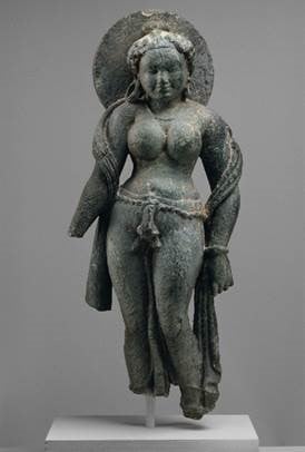 A bronze sculpture of a female figure, standing in a relaxed pose with one hip slightly raised. She is adorned with intricate jewelry, including earrings and a belt. Her hair is styled and gathered on top of her head, with a halo-like circular element behind it. The sculpture is detailed, with defined features and a graceful, flowing drapery.