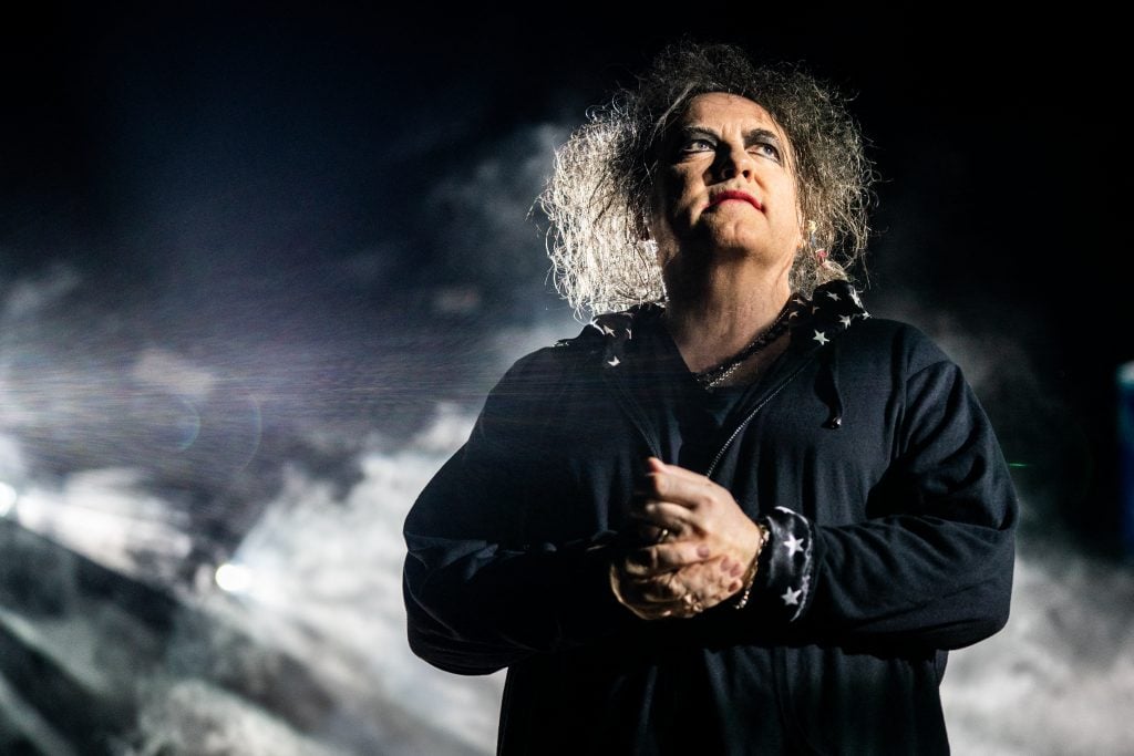 The Cure frontman standing on a dark stage, with light and smoke behind him