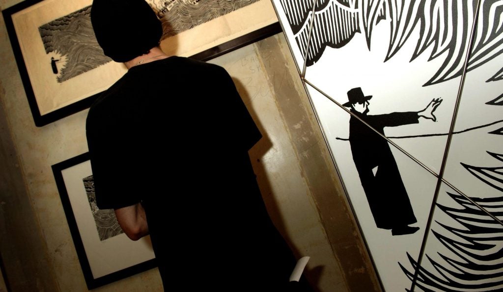 man in a black shirt with short black hair standing in front of a wall covered with framed drawings of black and white pictures