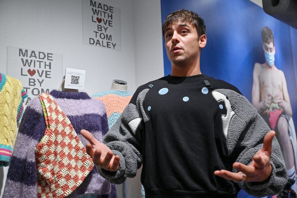 Diver Tom Daley in a sweater gesturing while standing next to mannequins dressed in his knitted designs