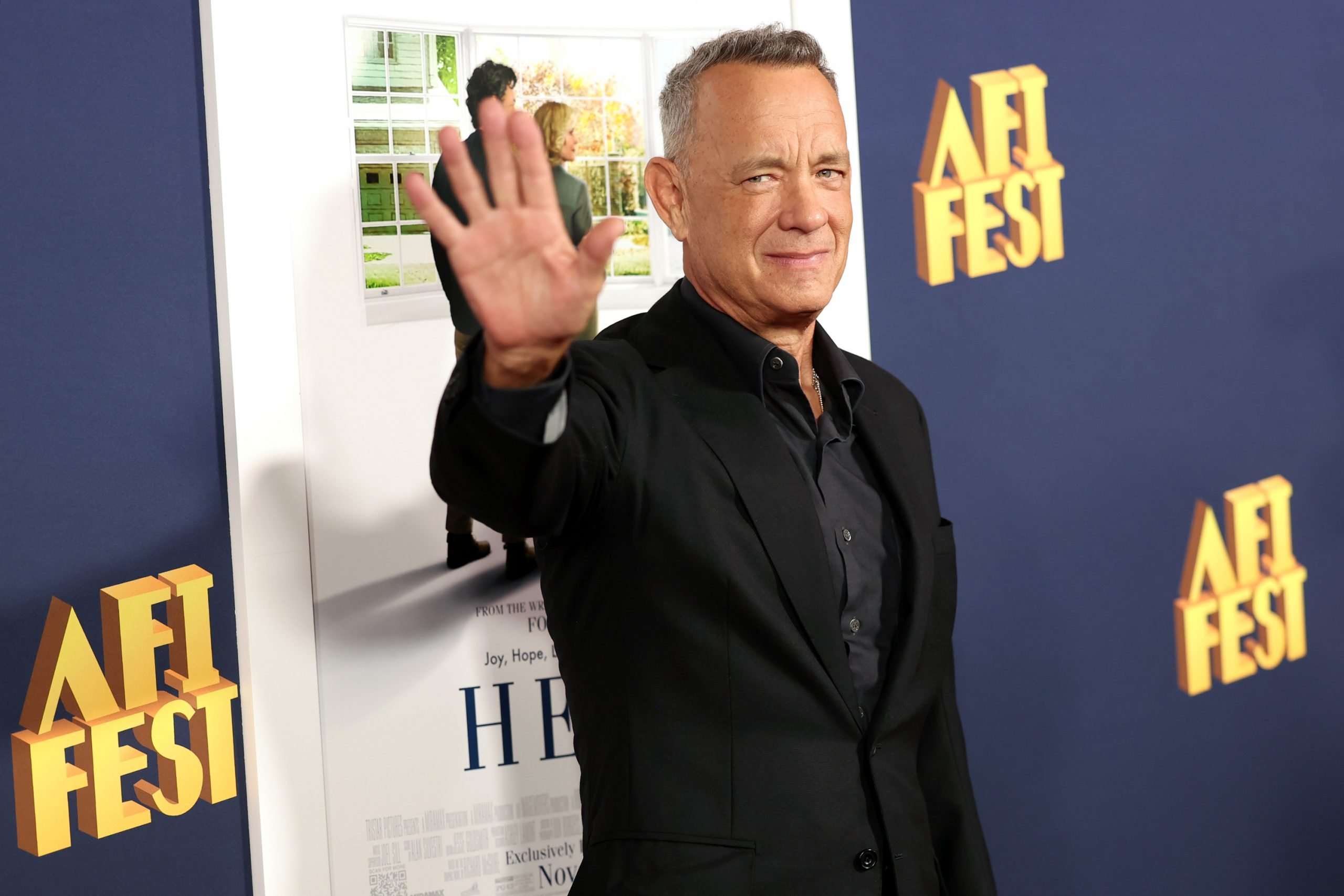 A photograph of Tom Hanks waving in an all black suit on the red carpet of a film premier.