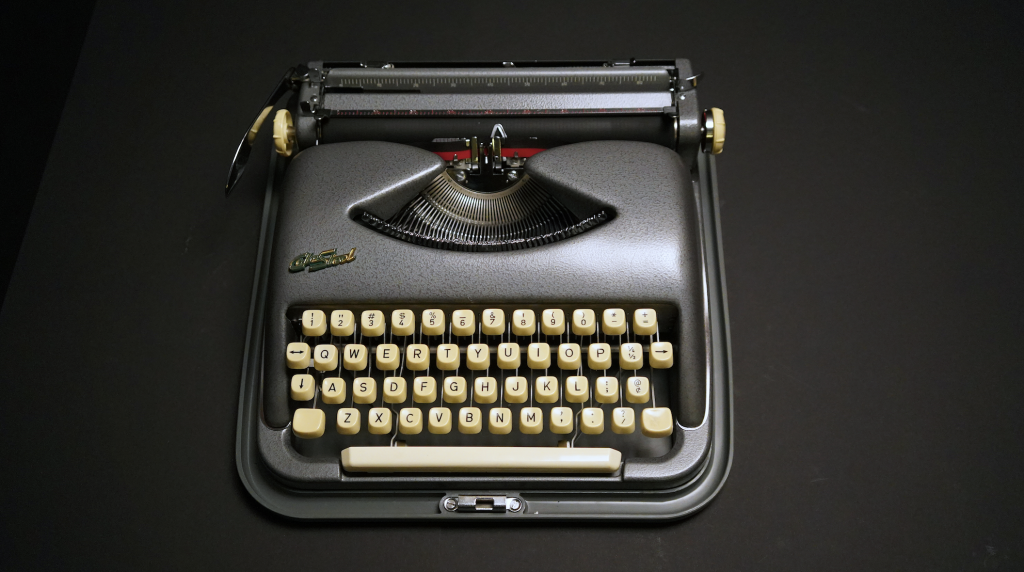 A photograph taken from above of a vintage grey typewriter, on a black background.