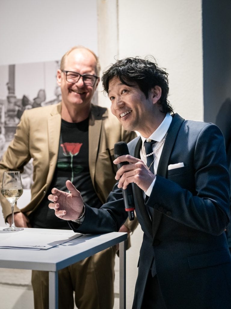 Artist Tomokazu Matsuyama wearing a navy suit speaking animatedly into a microphone behind which is and curator Cristoph Doswald wearing a beige blazer and graphic tee behind him smiling.