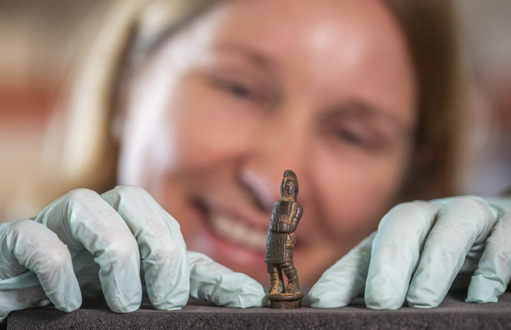 A woman wearing plastic gloves while handling a tiny sculpture of a gladiator