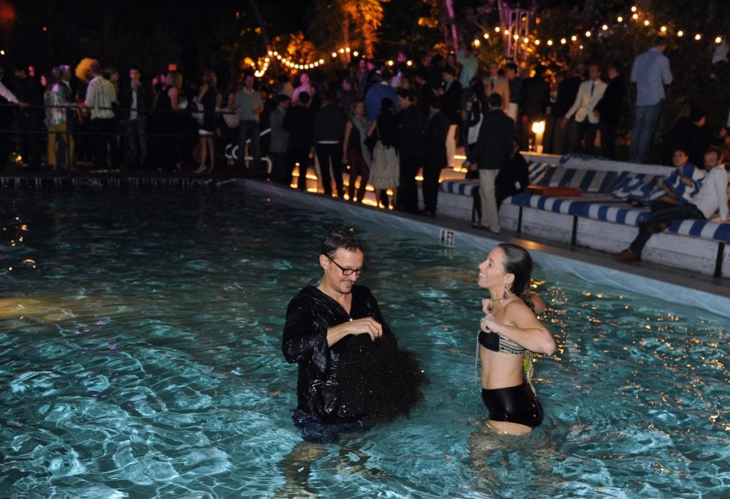 A lively scene at a nighttime poolside party, with many people mingling in the background under string lights and trees. In the pool, a man in glasses and a dark shirt, visibly wet, smiles as he stands chest-deep in the water. Beside him, a woman in a black and tan two-piece swimsuit, with long earrings and her hair slicked back, appears to be adjusting her top while smiling at him