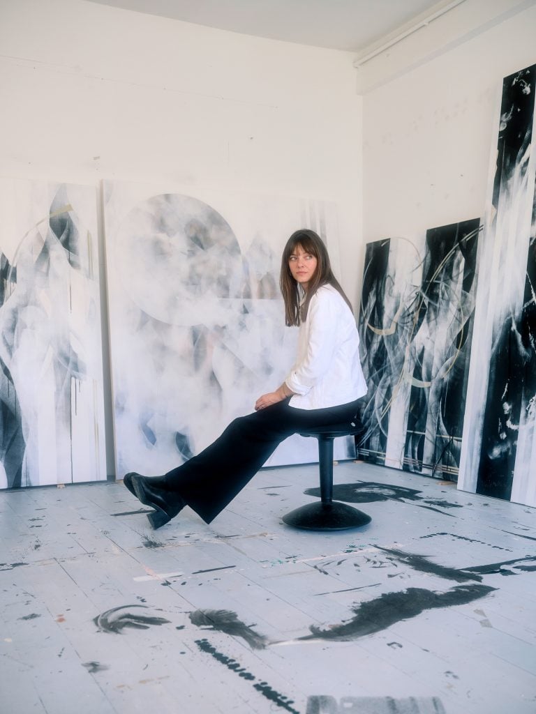 a woman named Ruscha Voormann sits on a stood amid her black and white abstract paintings