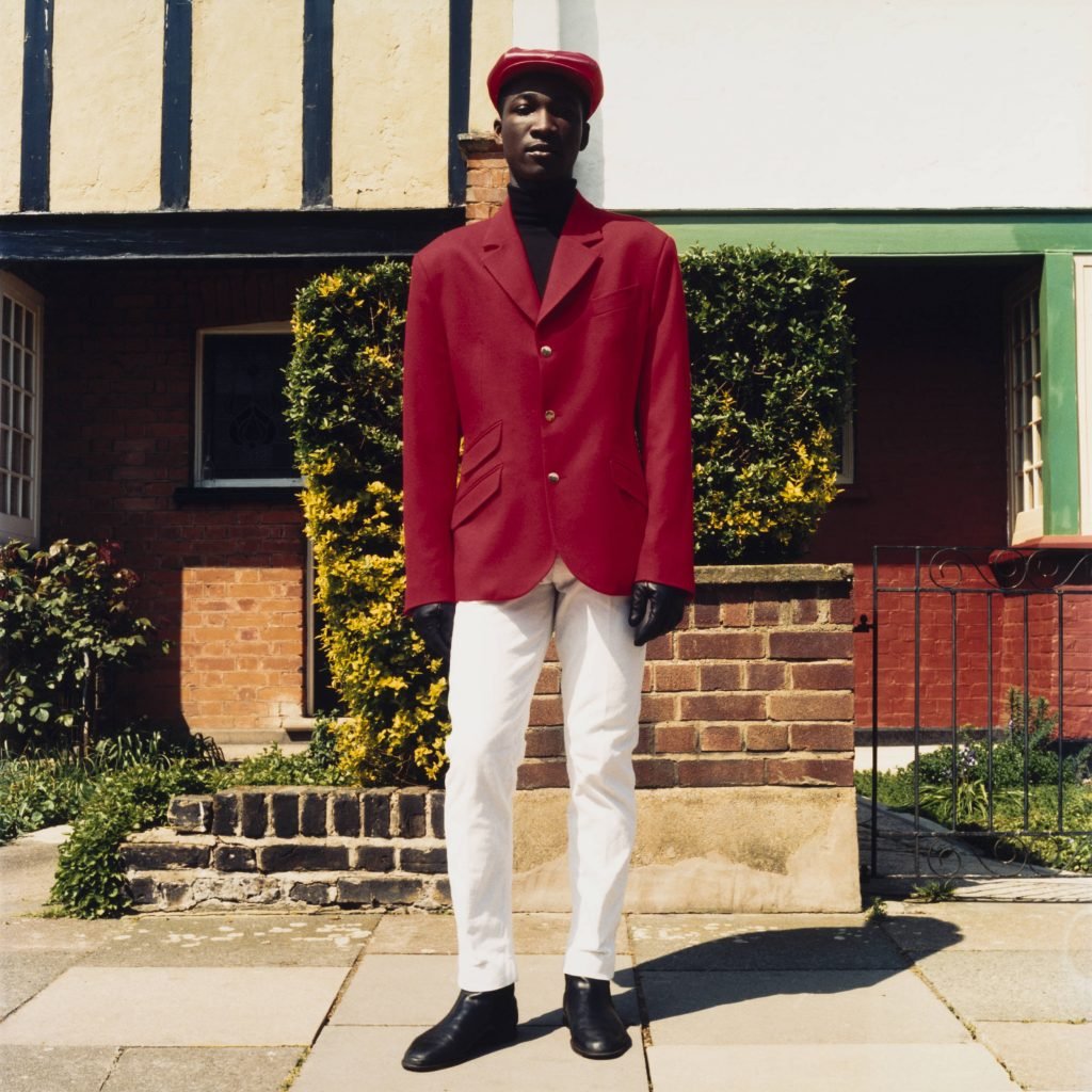 This photo depicts a black man sporting a hat, a red suit jacket and white pants, black shoes standing against a brick wall