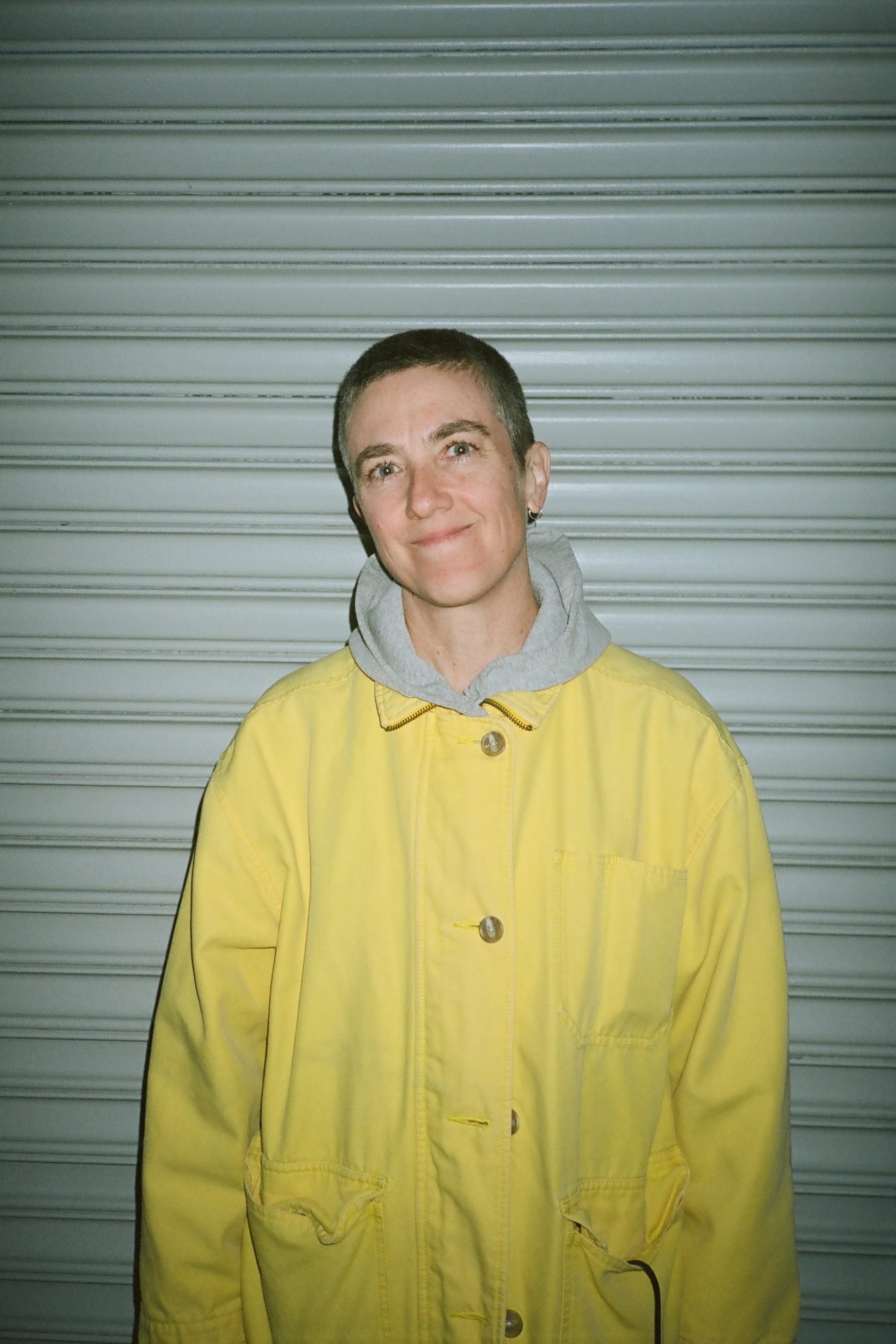 a person in a yellow car with a grey hooded sweatshirt and bright eyes stands smiling in front of a door grate