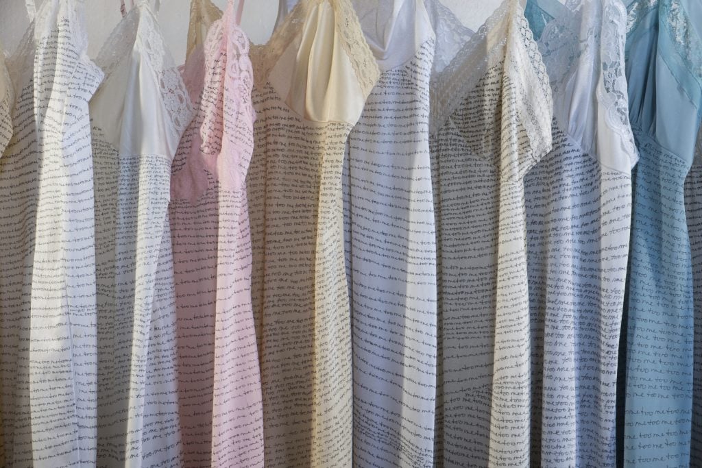 A photograph of delicate, vintage-style dresses hanging in a row, each featuring intricate lace details. The fabric of the dresses is covered with handwritten text that repeats the phrase "me too," creating a powerful visual statement. The dresses vary in soft pastel shades, including white, cream, pink, and blue, contrasting with the repeated words, evoking themes of vulnerability and resilience.