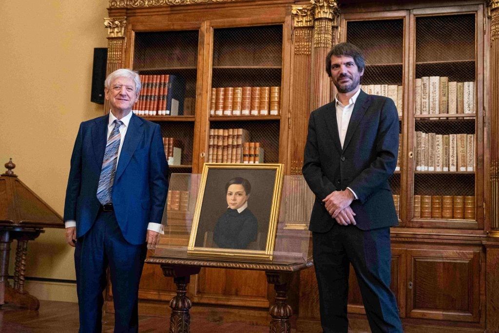 Two men stand next to a painting of a small spanish boy in a gold frame in a ceremony in a library