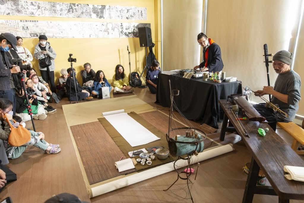 A musician on the DJ deck performing with another musician playing Er-hu in front of a crowd sitting on the floor. Most of them are East Asians.