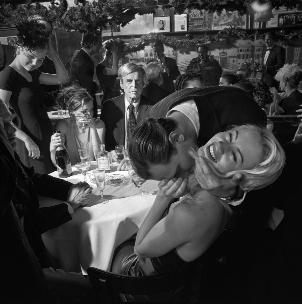 A black-and-white photograph of a lively social gathering, featuring a man playfully embracing a smiling woman in the foreground. A table with glasses, bottles, and elegantly dressed individuals surrounds them. In the background, a serious-looking man stares directly at the camera amidst a bustling crowd, creating a contrast to the lighthearted mood in the foreground.