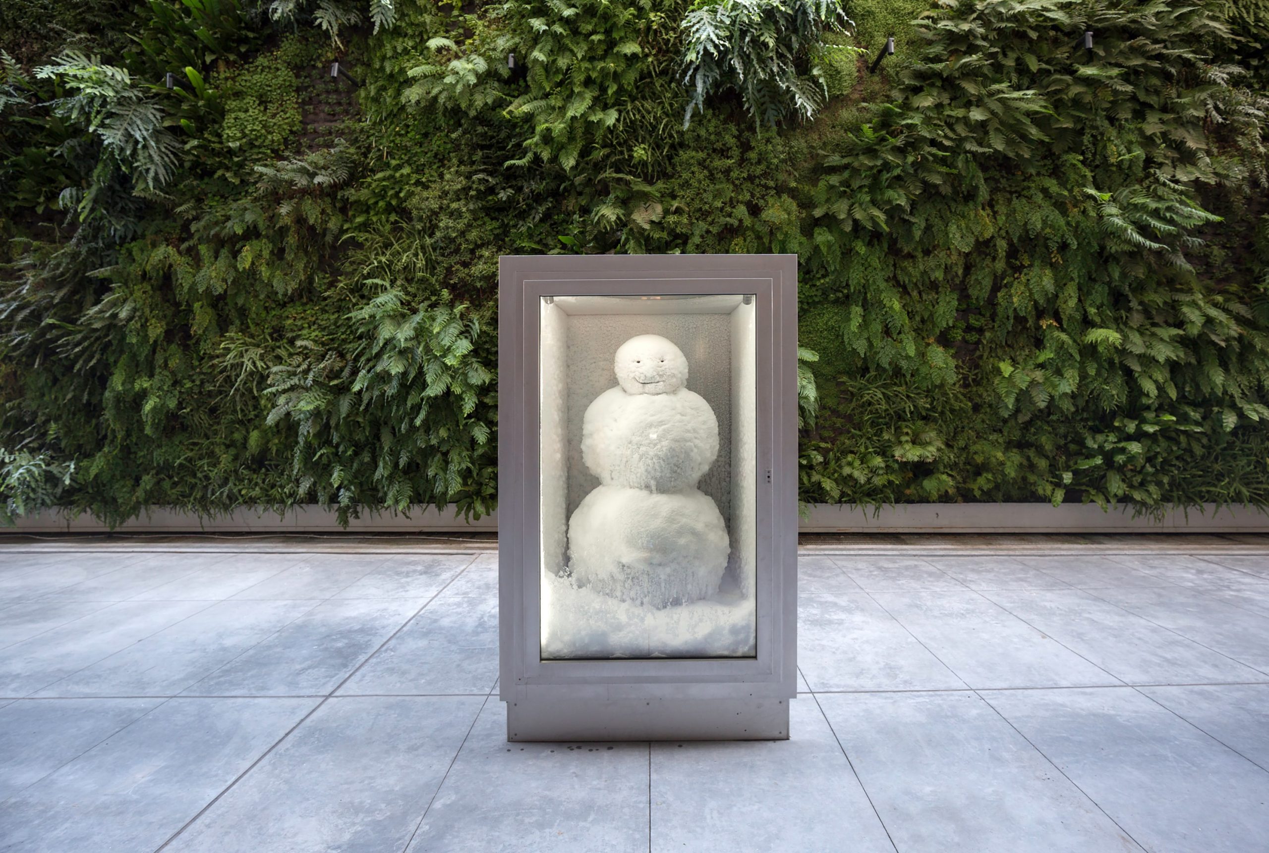 a snowman is in a refrigerated box next to a bush on a sunny day