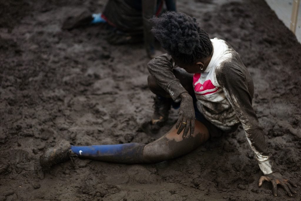 A performer sits on muddy ground, wearing a dirt-streaked white shirt and blue socks.