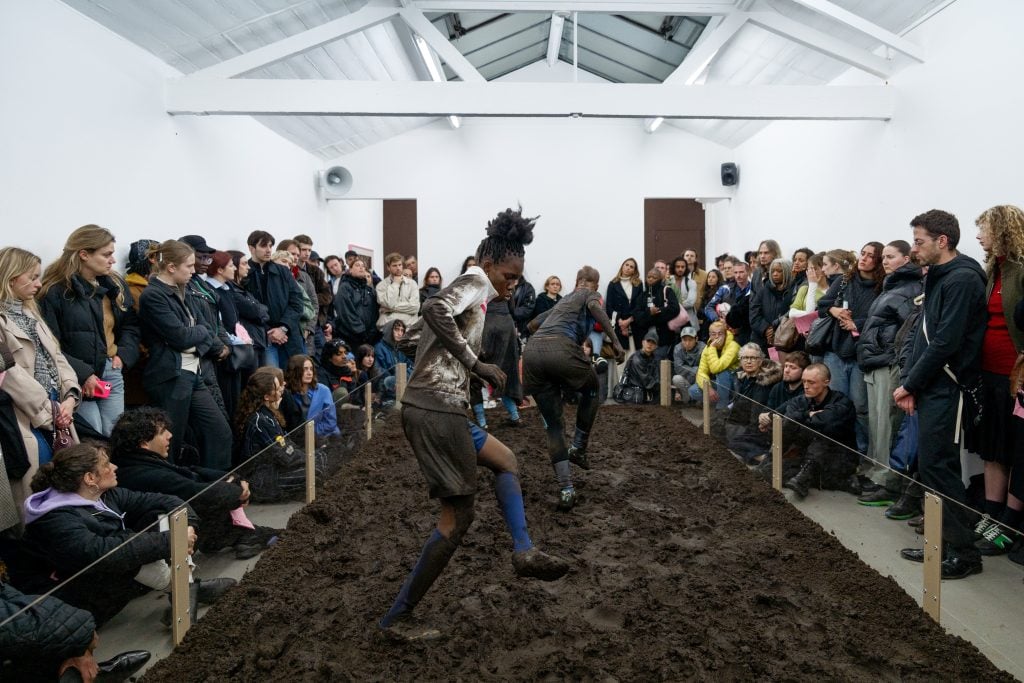 Performers engage in a dynamic mud-filled performance, surrounded by a captivated audience in an enclosed space.