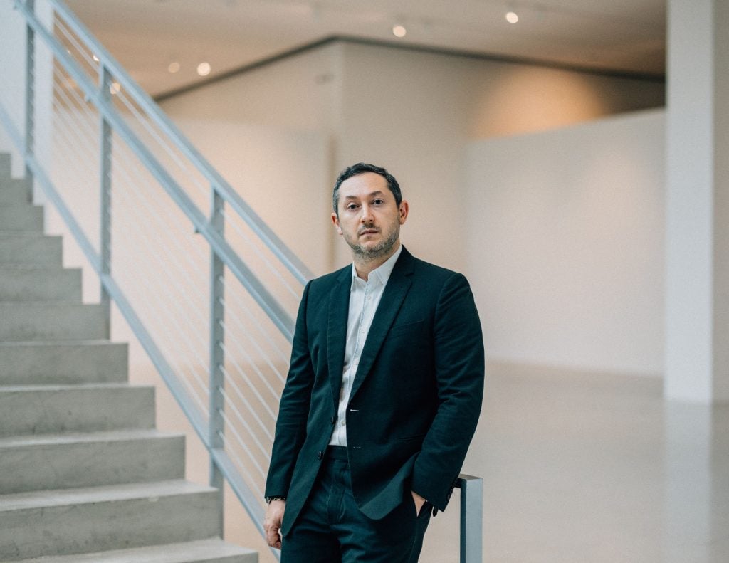 a man in a black suit and blue shirt with no tie stands inside of a stark modern building next to a stairwell