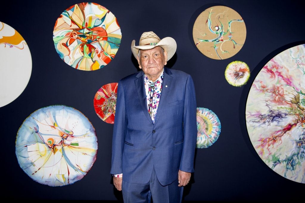 Alex Janvier, Indigenous artist, on the occasion of his career retrospective at the McMichael Canadian Art Collection in Kleinburg. His is an elderly man in a blue suit with a colorful tie and a tan cowboy hat, standing in front of a black gallery wall hung with an array of medium-sized abstract circular canvases. 