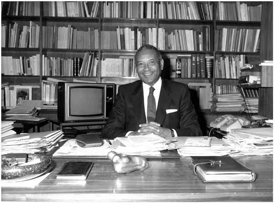 A black and white photo of Amadou-Mahtar M’Bow sitting at an office desk in front of a wall-to-wall bookcase. 