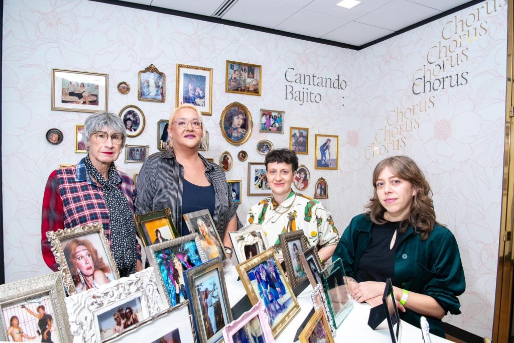 Four people standing in an artwork installation with various framed archival photographs standing upright and on the wall behind them that reads “Cantando Bajito: Chorus”