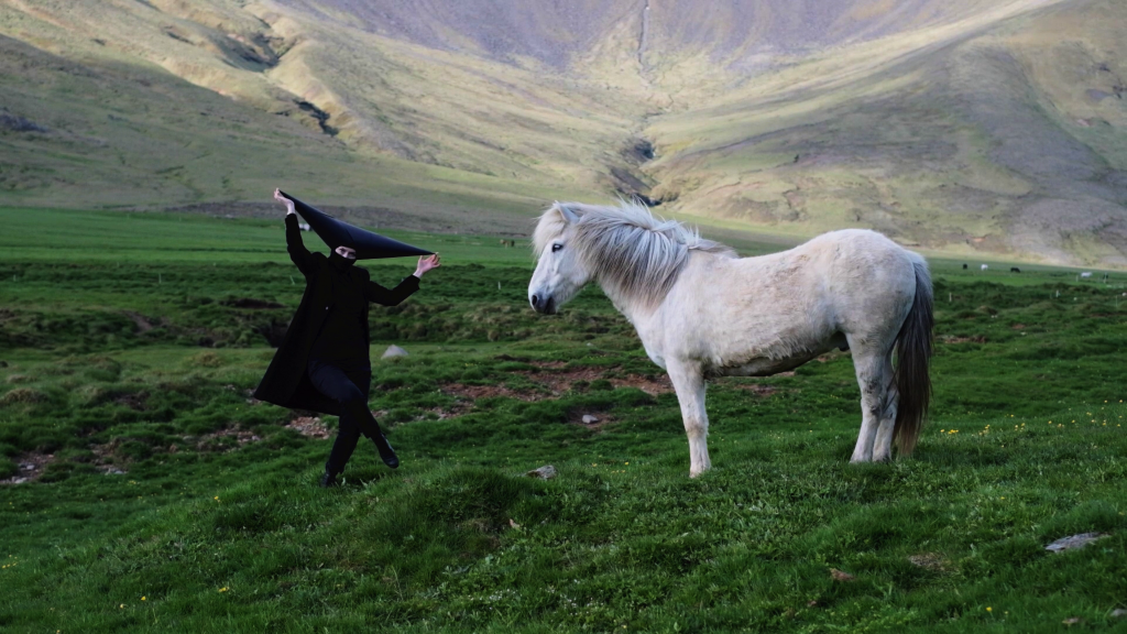 Photo of a woman in a black dress dancing on green grass land, her hands up holding the tip of her headpiece which is in the shape of an inverted triangle. She is posing next to a white horse.