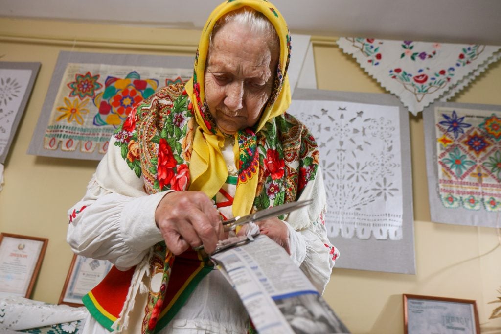 an old woman in traditional dress uses scissors to cut a hunk of newspaper, there are patterns cut into paper hanging on the wall behind