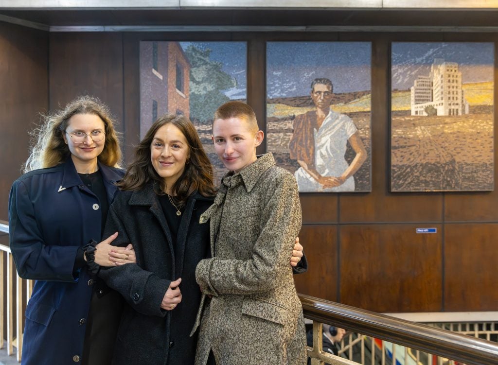 Three women posing in front of a mural in a train station