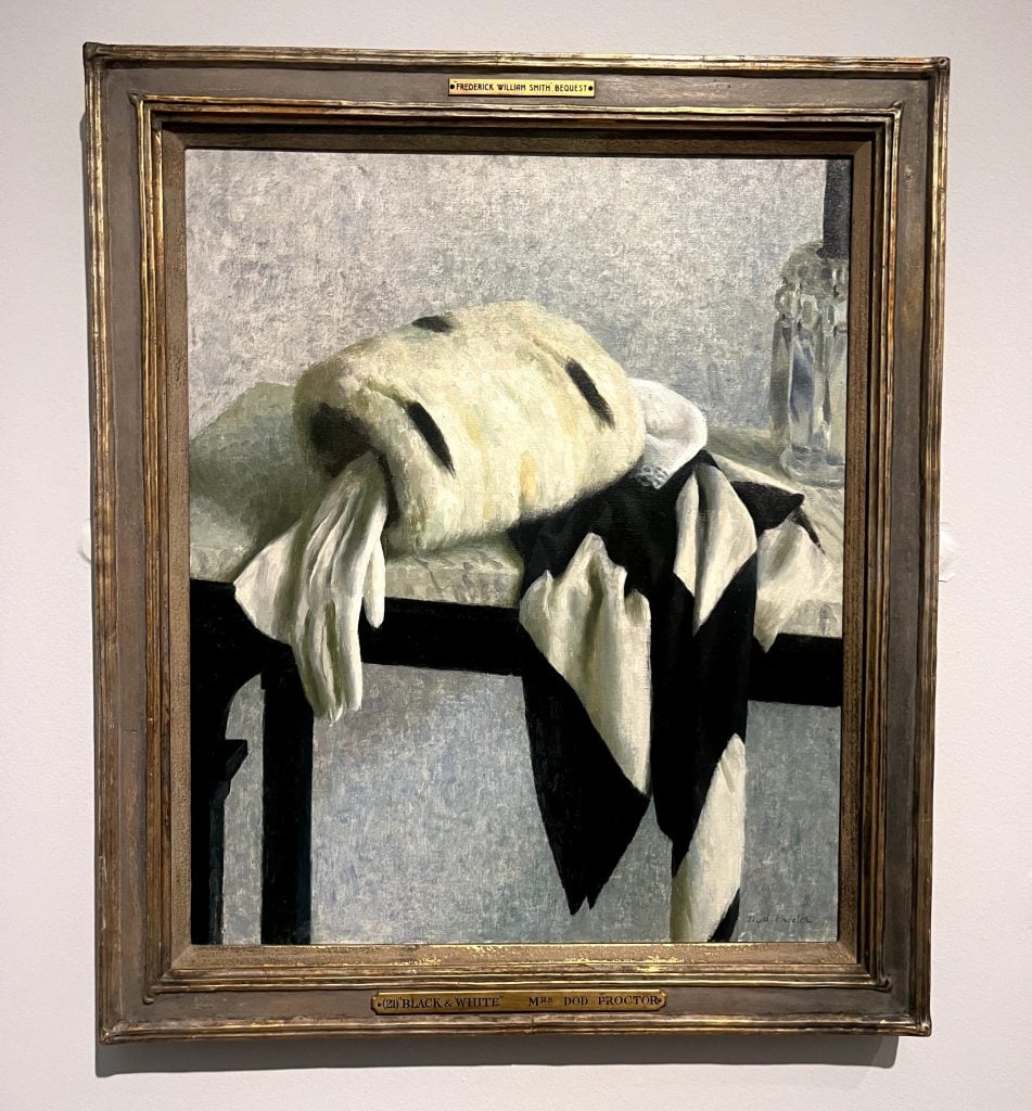 a photograph of a framed painting hanging on the wall and the painting contains a monochromatic arrangement of a muff with a silk scarf and white glove hanging down over the side of a white marble side table, there is part of a crystal cut glass to the right just going off the frame, the walls are white