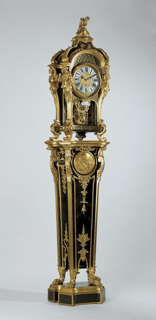 A gold and dark wood pedestal clock against a grey background