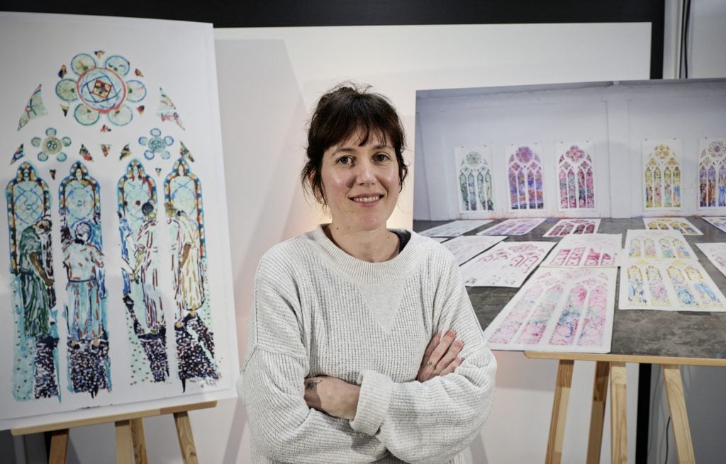 French artist Claire Tabouret, a young dark-haired white woman in a white shirt, stands in front of photos of her designs for new stained glass windows for Notre Dame Cathedral in Paris.