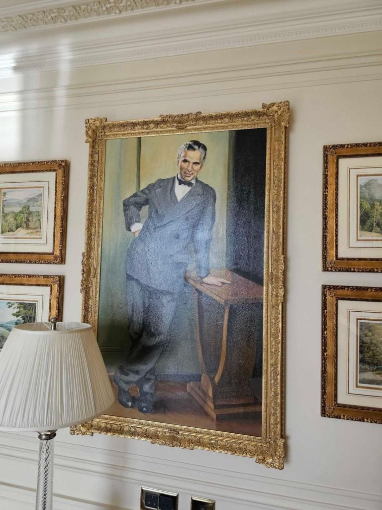 photograph of a large portrait of a young man in a black suit leaning against a table surrounded by a handcarved frame alongside other smaller pictures on a hotel room wall