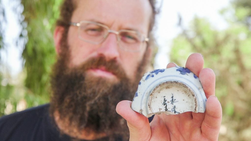 A bearded man holding up a broken pottery shard with Mandarin characters inscribed at its base