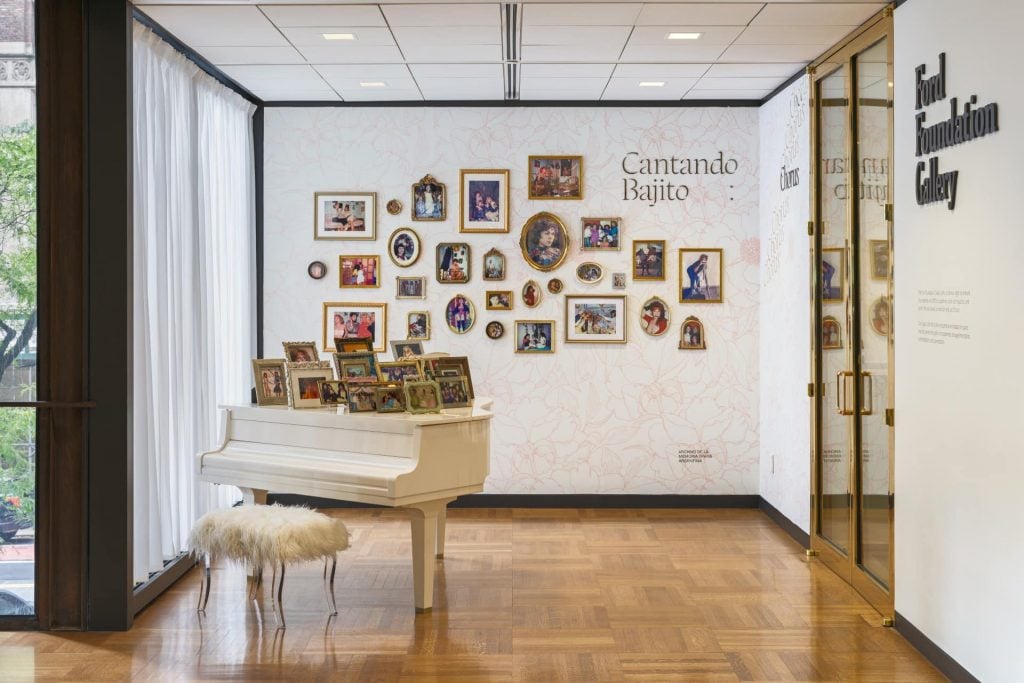 Installation of a grand piano with framed photos on the lid and on the wall behind it in front of a gallery entrance. 