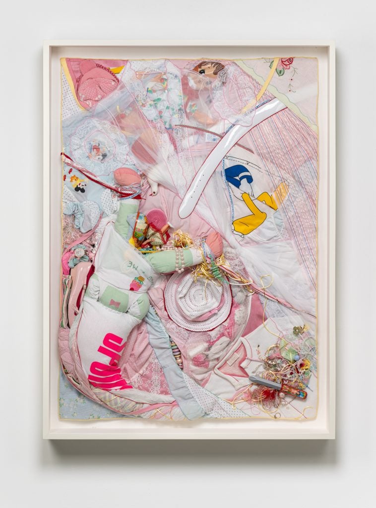 an assemblage artwork with a soft plush doll with splayed legs, a shirt with the word Spoiled written on it, and other pink fabrics and knicknacks