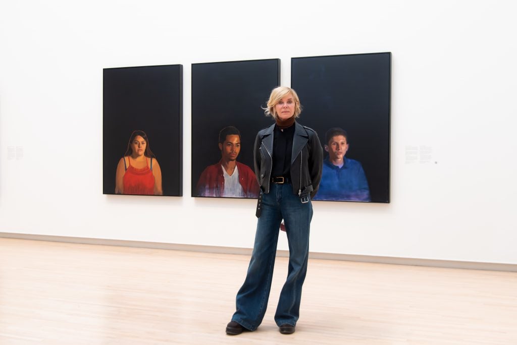 a blonde woman clad in black stands in a museum gallery in front of images of paintings of three children with black backgrounds