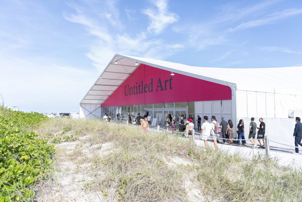 People lining up to get into a large white tent located next to a grassland under a blue sky. At the entrance of the tent sees a fuchsia banner which has "Untitled Art" spelt out.