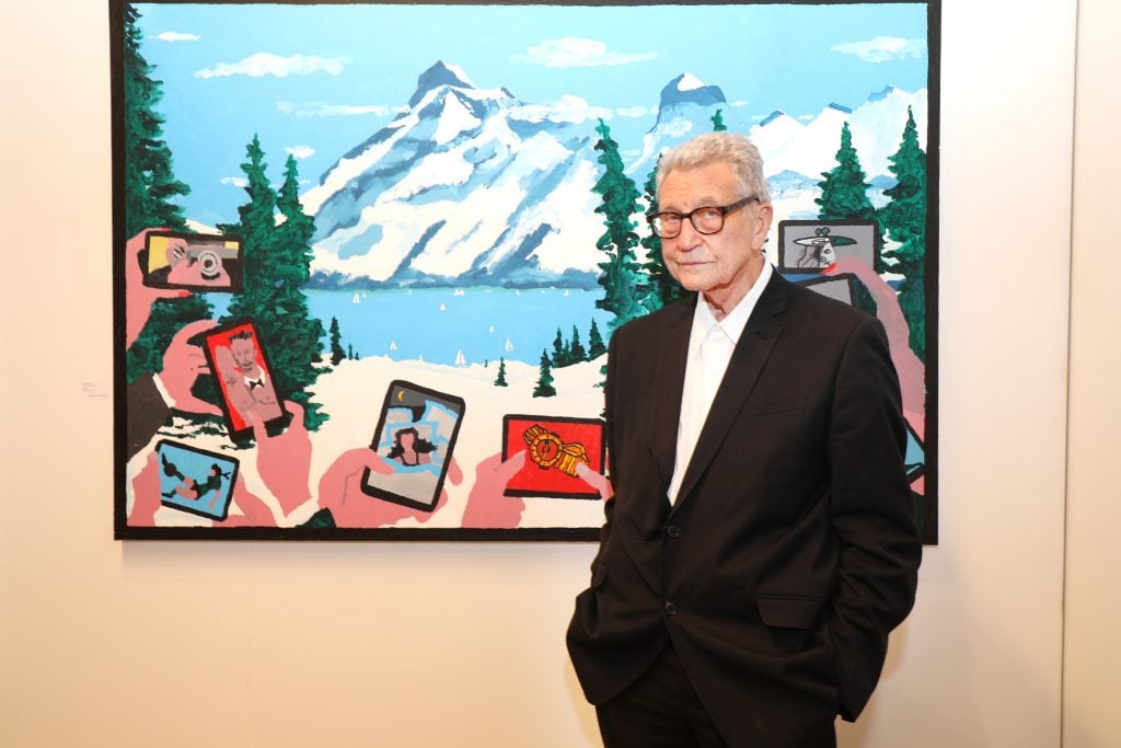 A colorful photograph of artist Derek Boshier as an old man, wearing a black suit, standing in front of a large painting of a vista of mountain and a lake, with hands holding up smartphones to photograph the view in the foreground. 