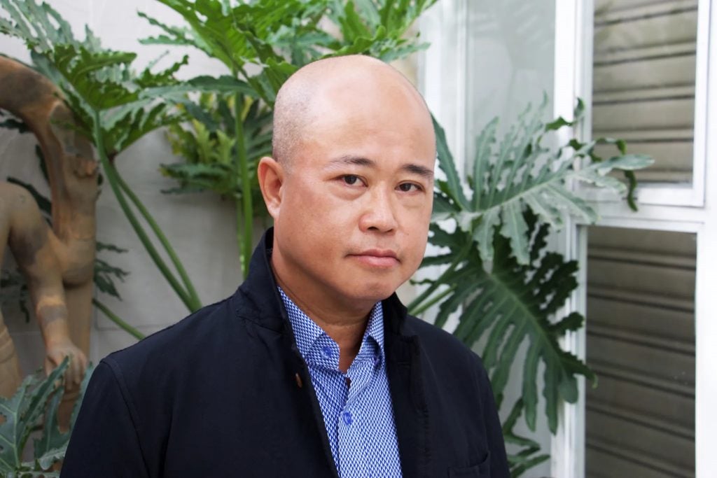 A color photo of a middle-aged bald Asian man in a blue suit jacket and blue colored shirt, seen from the shoulders up in front of a palm-like plant with large leaves. 