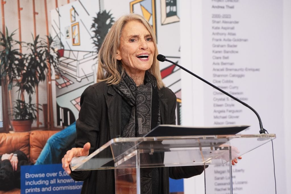 Dorothy Lichtenstein at the Roy Lichtenstein Foundation's 100th birthday party for the late artist. She is an elderly woman with shoulder length ash blonde hair, standing at a podium with a microphone in front of a wall covered with a large comic book strip-style artwork.