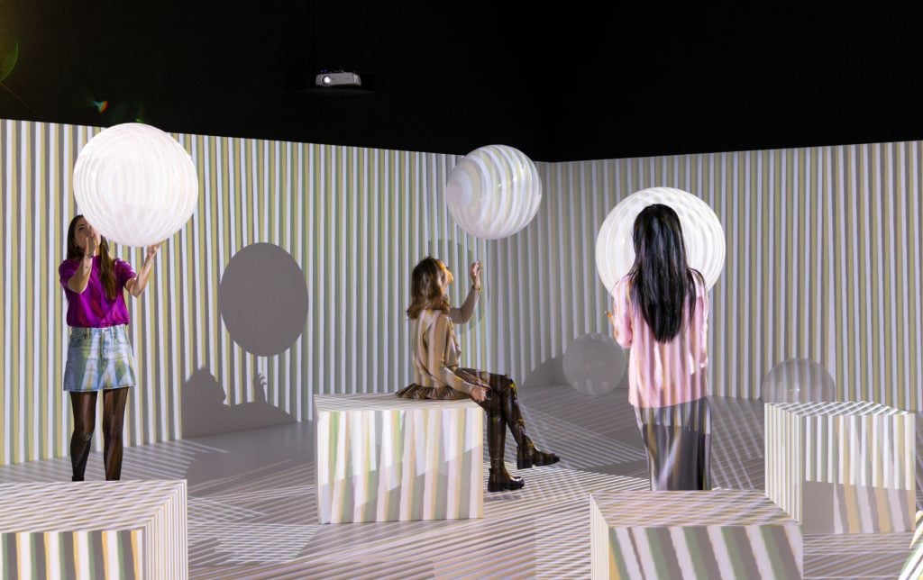 people mill and play in a room filled with bouncy balloon balls and squares to sit on, and projectors around the room beam stripes over the whole room