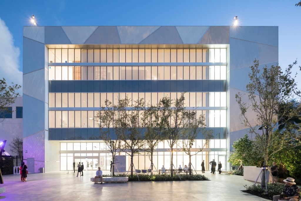 A modern building with a geometric facade and large illuminated windows, surrounded by trees and a paved courtyard at dusk