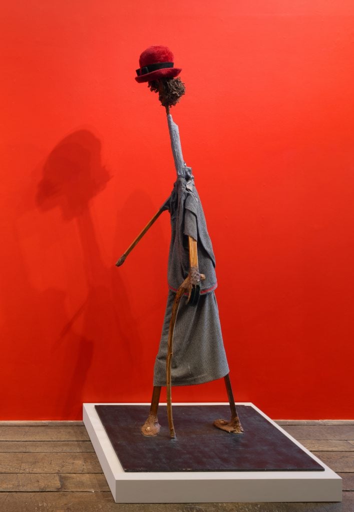 a thin sculpture with a small head set against a dark red wall