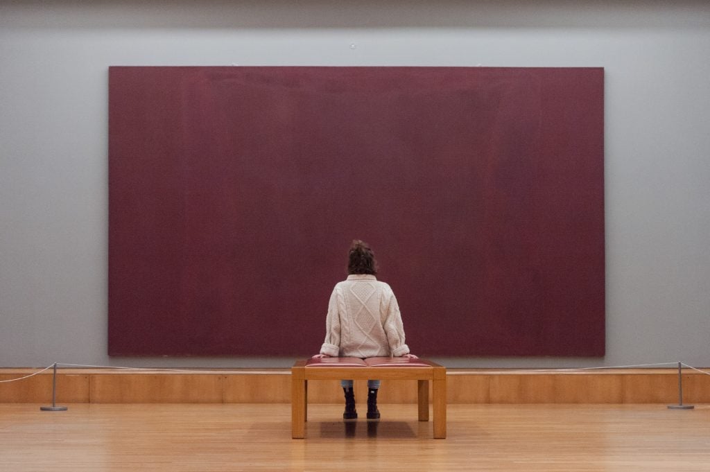 a woman sitting on a bench in front of a massive marroon painting is seen from behind