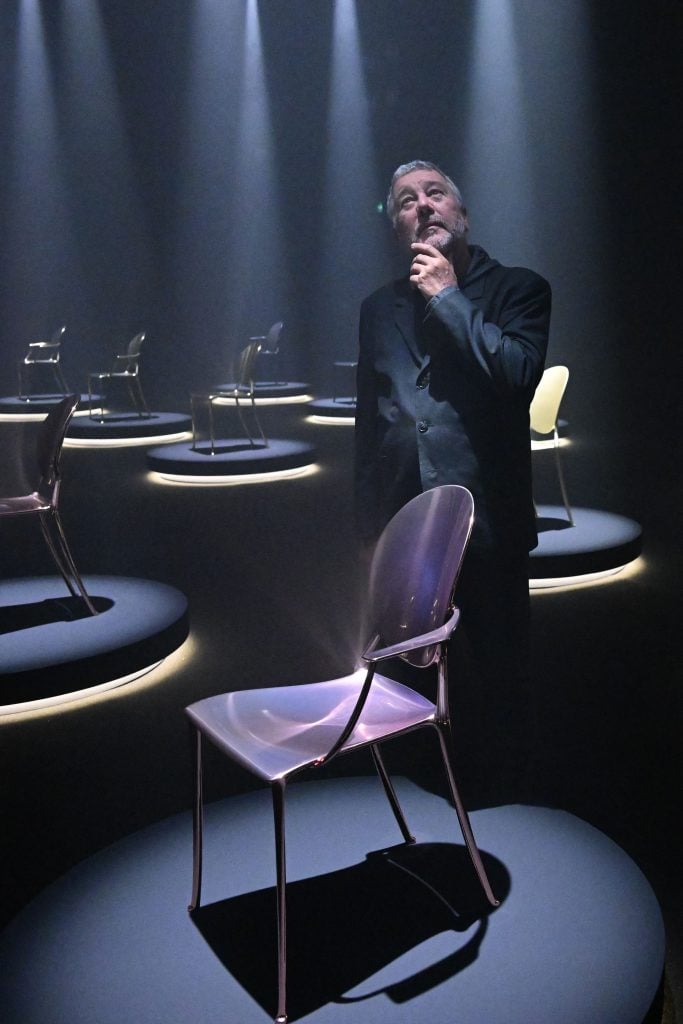 A man in a dark suit contemplates a sleek, translucent chair under dramatic spotlighting, surrounded by similarly illuminated chairs in a dim exhibition space.