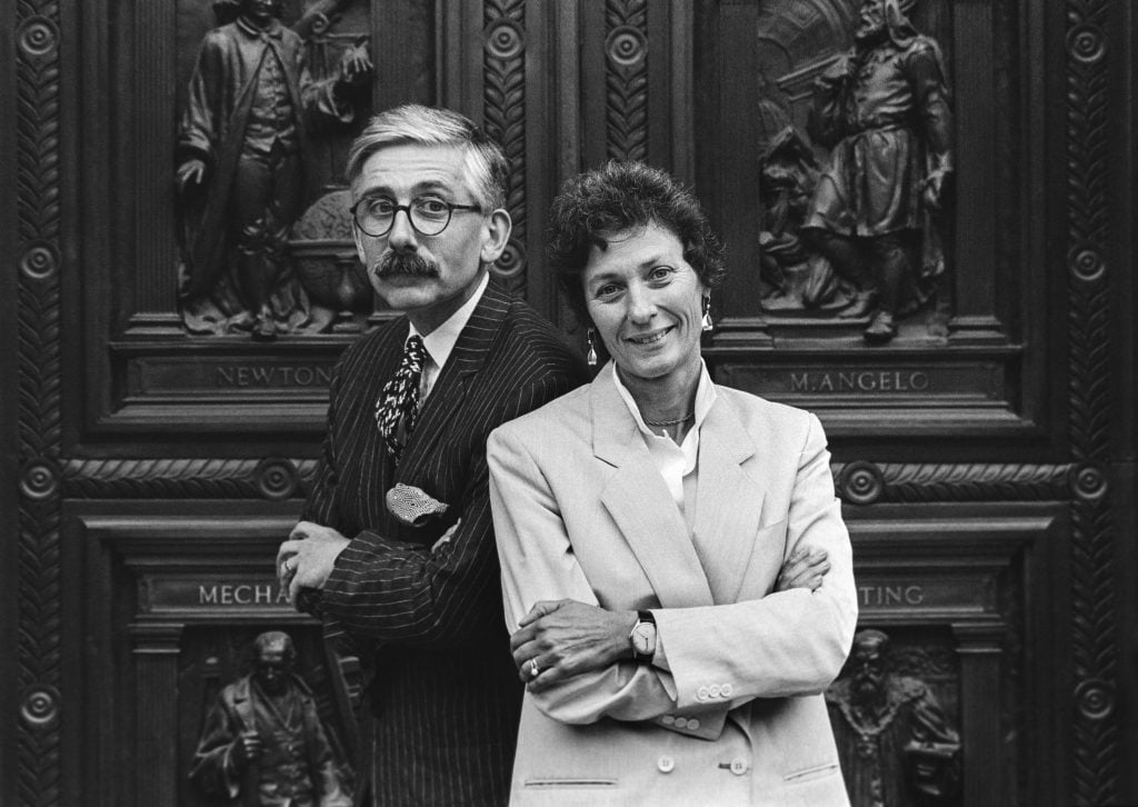 A black and white photo of a older white man with a mustache and thick rimmed glasses, and a younger white woman with short dark hair in light-colored power blazer, standing with their arms crossed in front of a massive, ornately carved wooden door. 